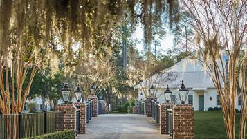 A paved pathway leading to a gated entrance with brick pillars and black metal railings, surrounded by trees with Spanish moss and a white building in the background.