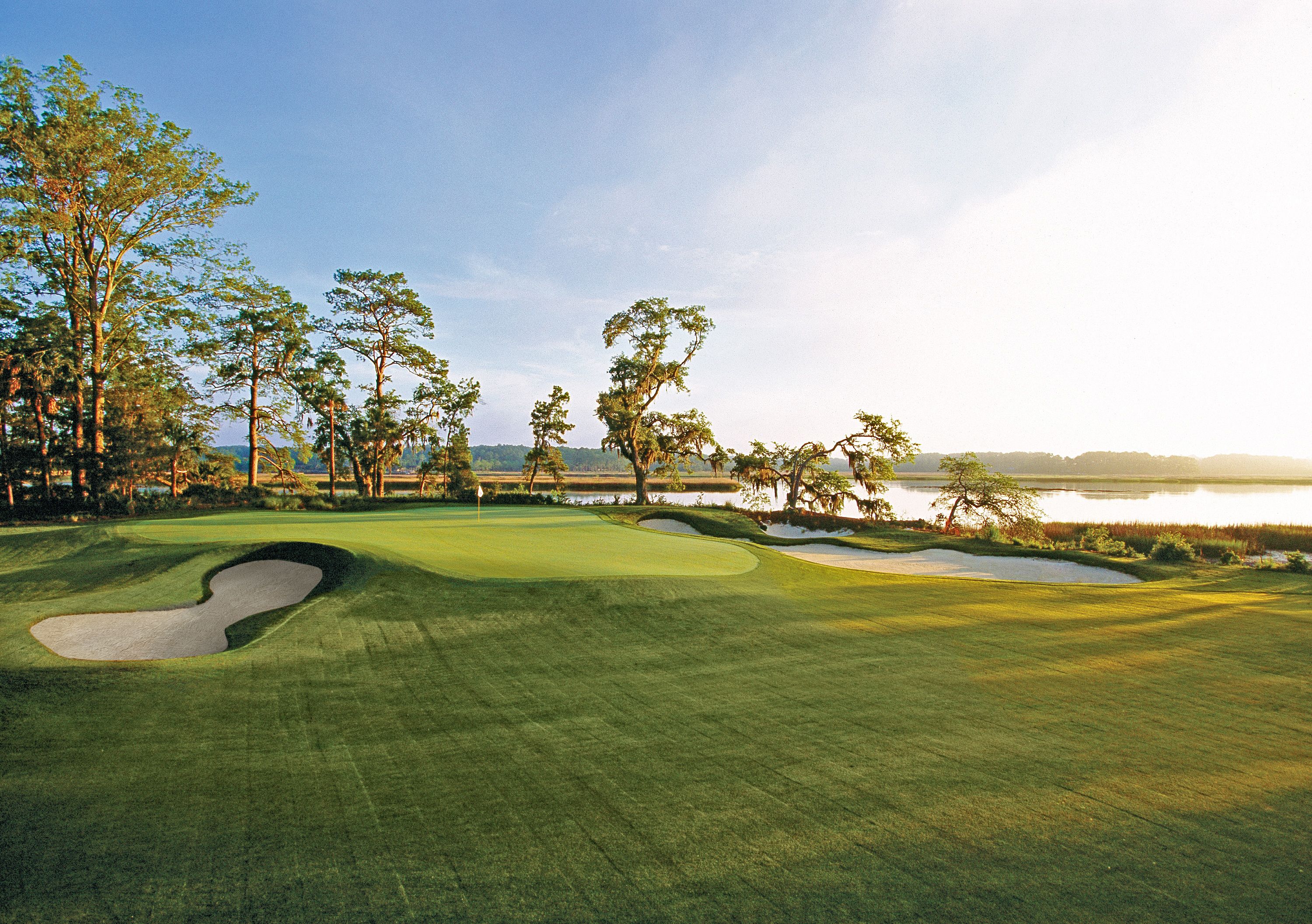 A golf course with sand bunkers, trees, and a water body in the background.