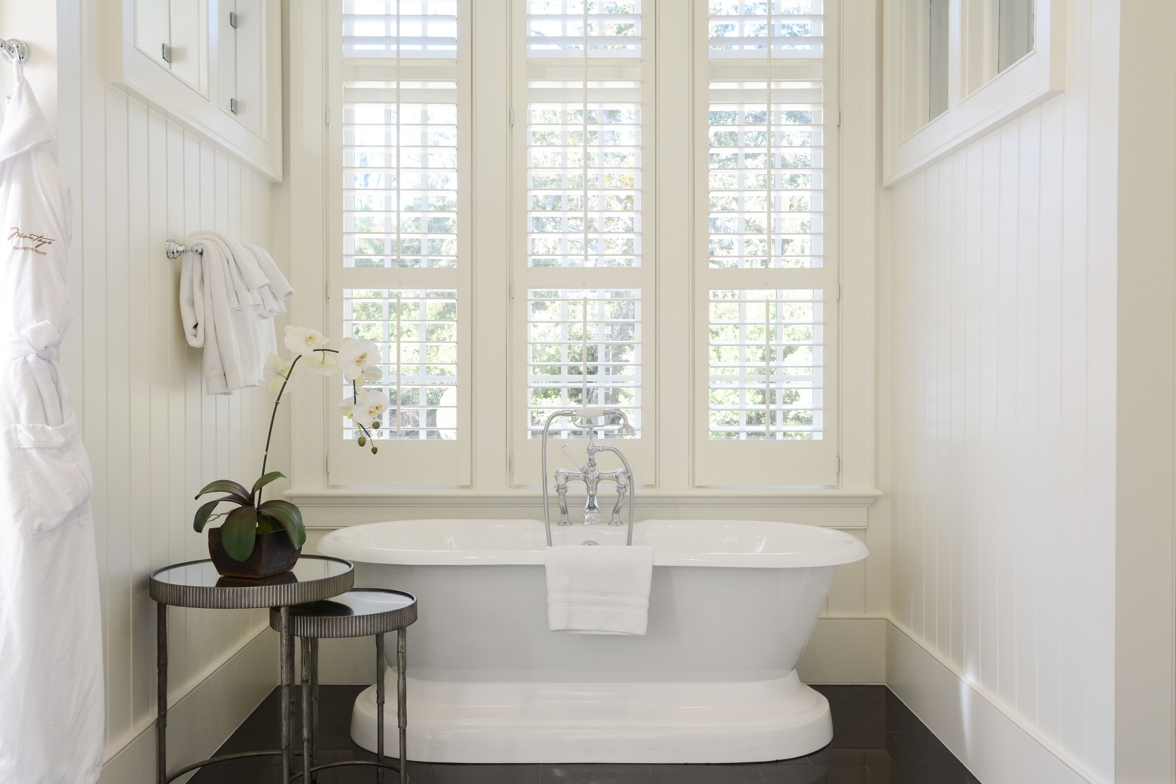 A bathroom with a freestanding bathtub, a small round table with a potted plant, and a window with blinds.