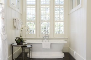 A bathroom with a freestanding bathtub, a small round table with a potted plant, and a window with blinds.