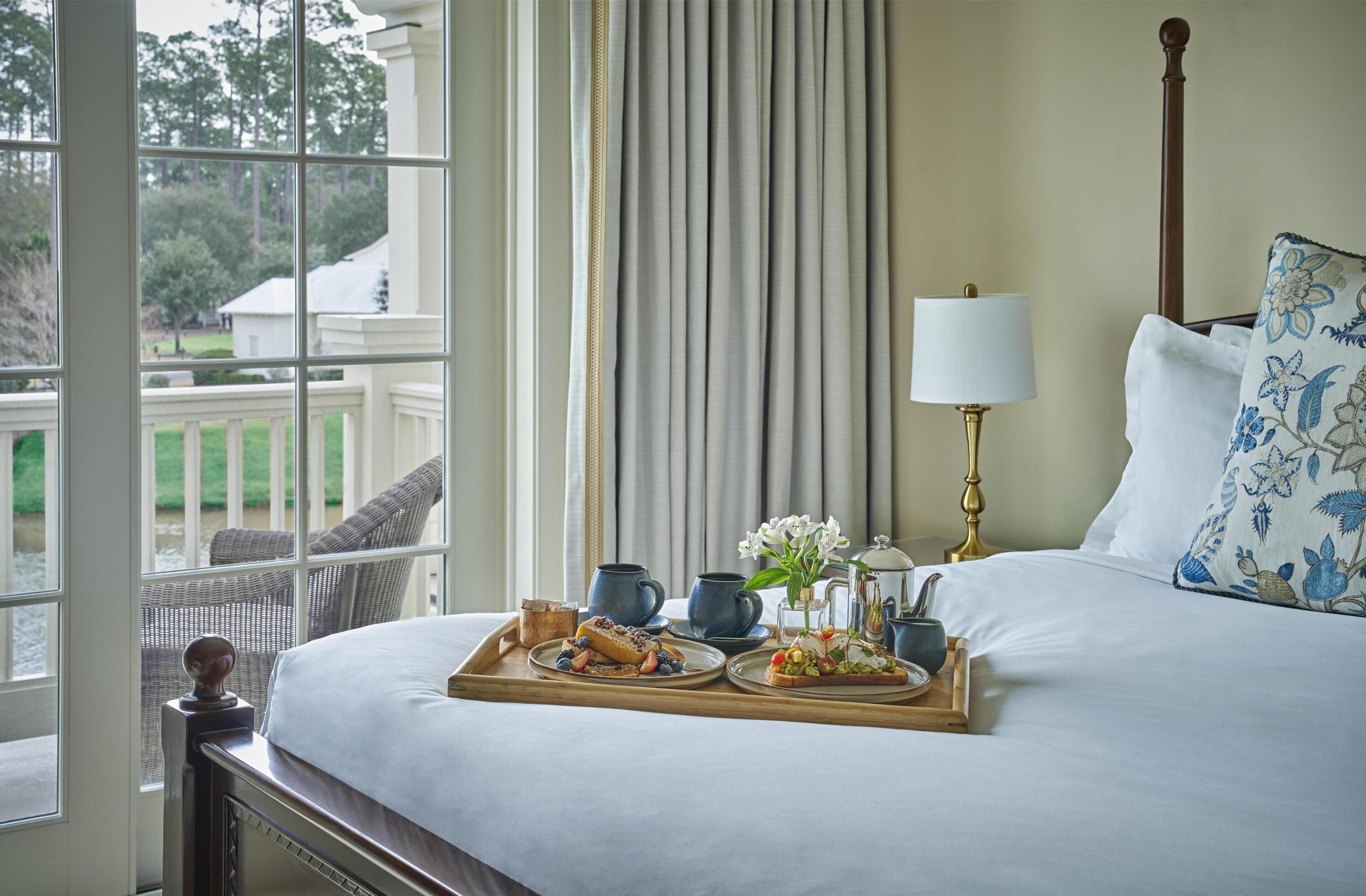 Una cama bien tendida con una bandeja de desayuno, una lámpara y vista a un jardín a través de una ventana.