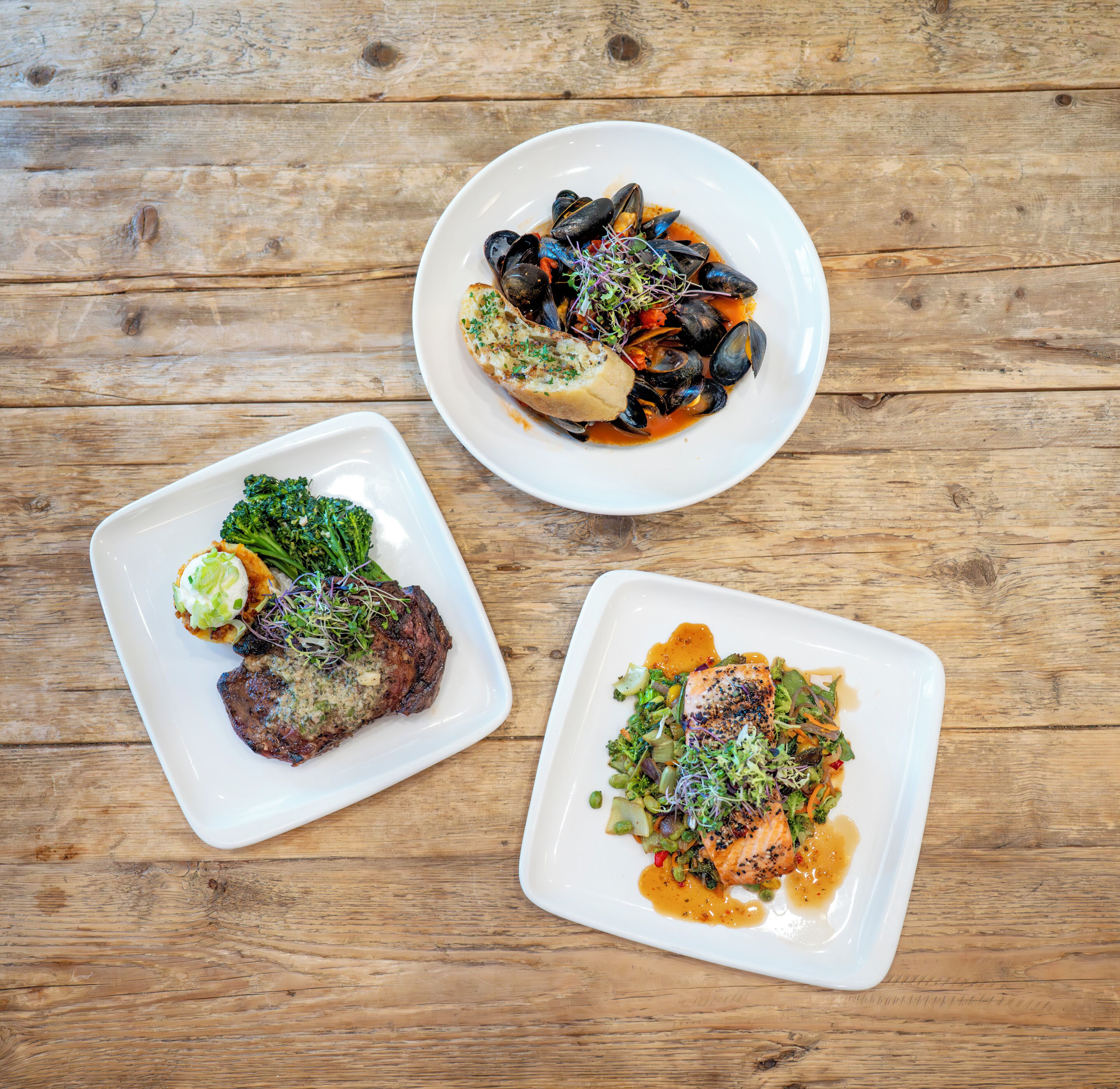 Three square white plates of food on a wooden table.