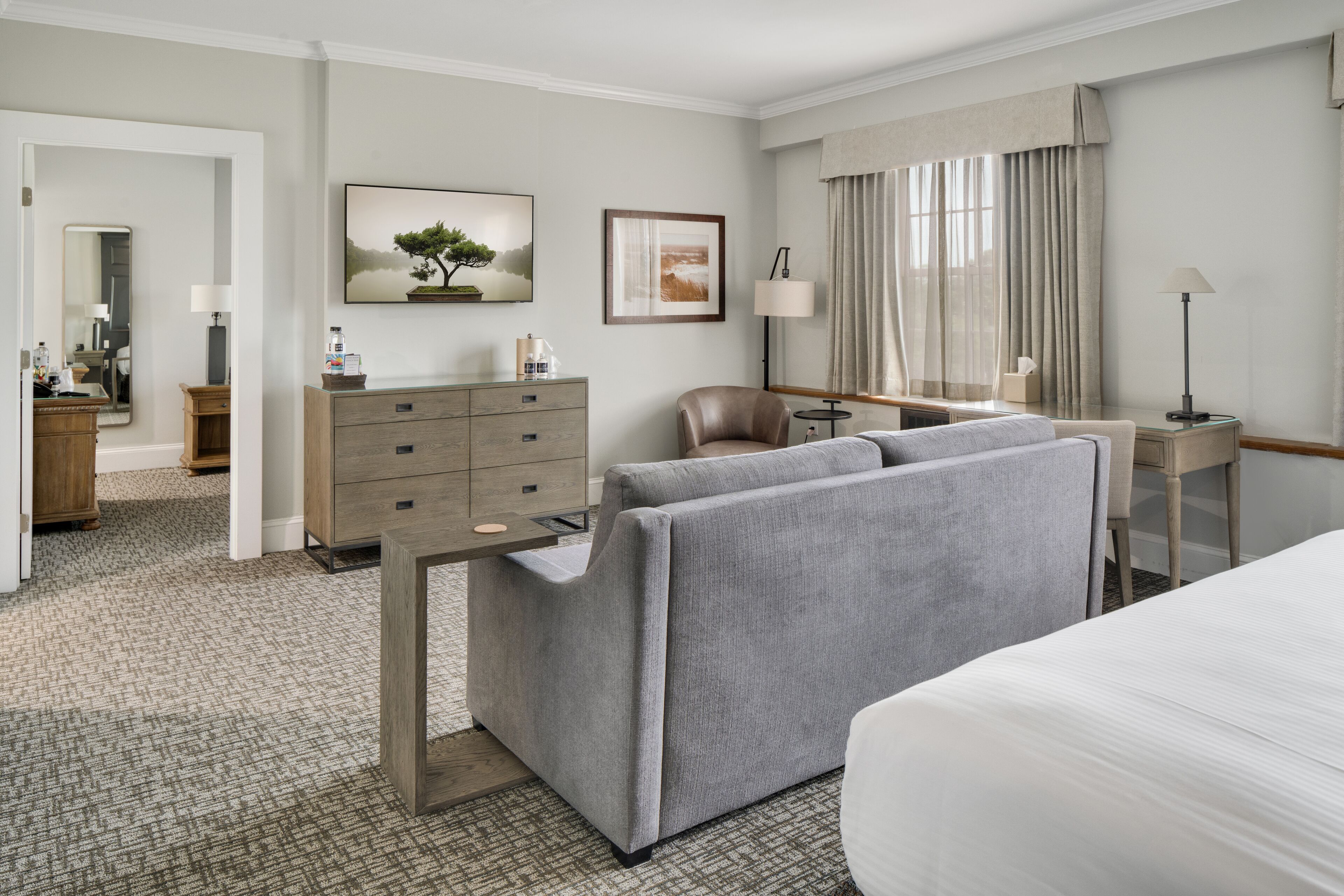A hotel room with a bed, a sofa, a desk, and a chair. There is a dresser with drawers and a mirror.