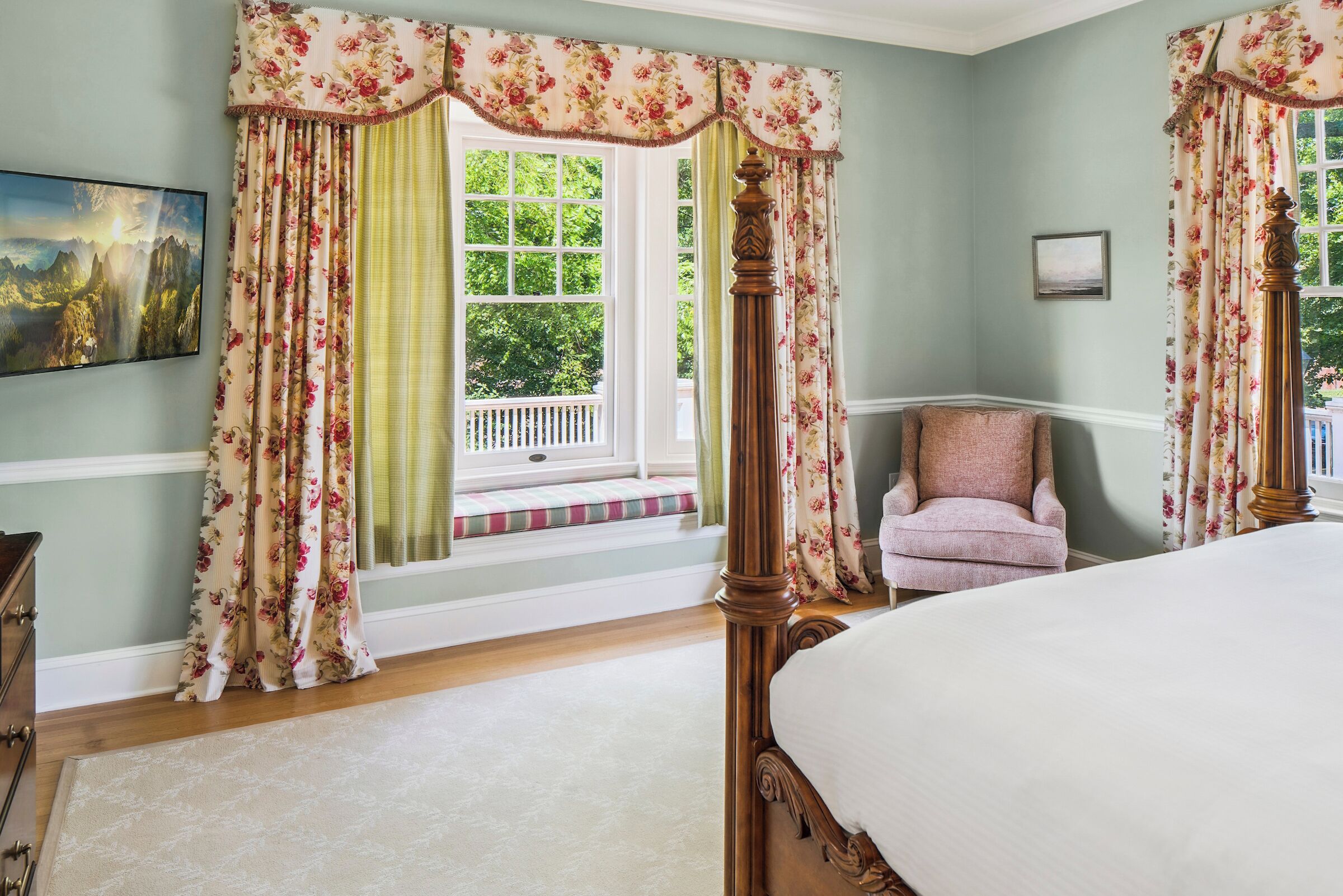 Un dormitorio con cama con dosel, cortinas florales, un banco junto a la ventana y una silla.