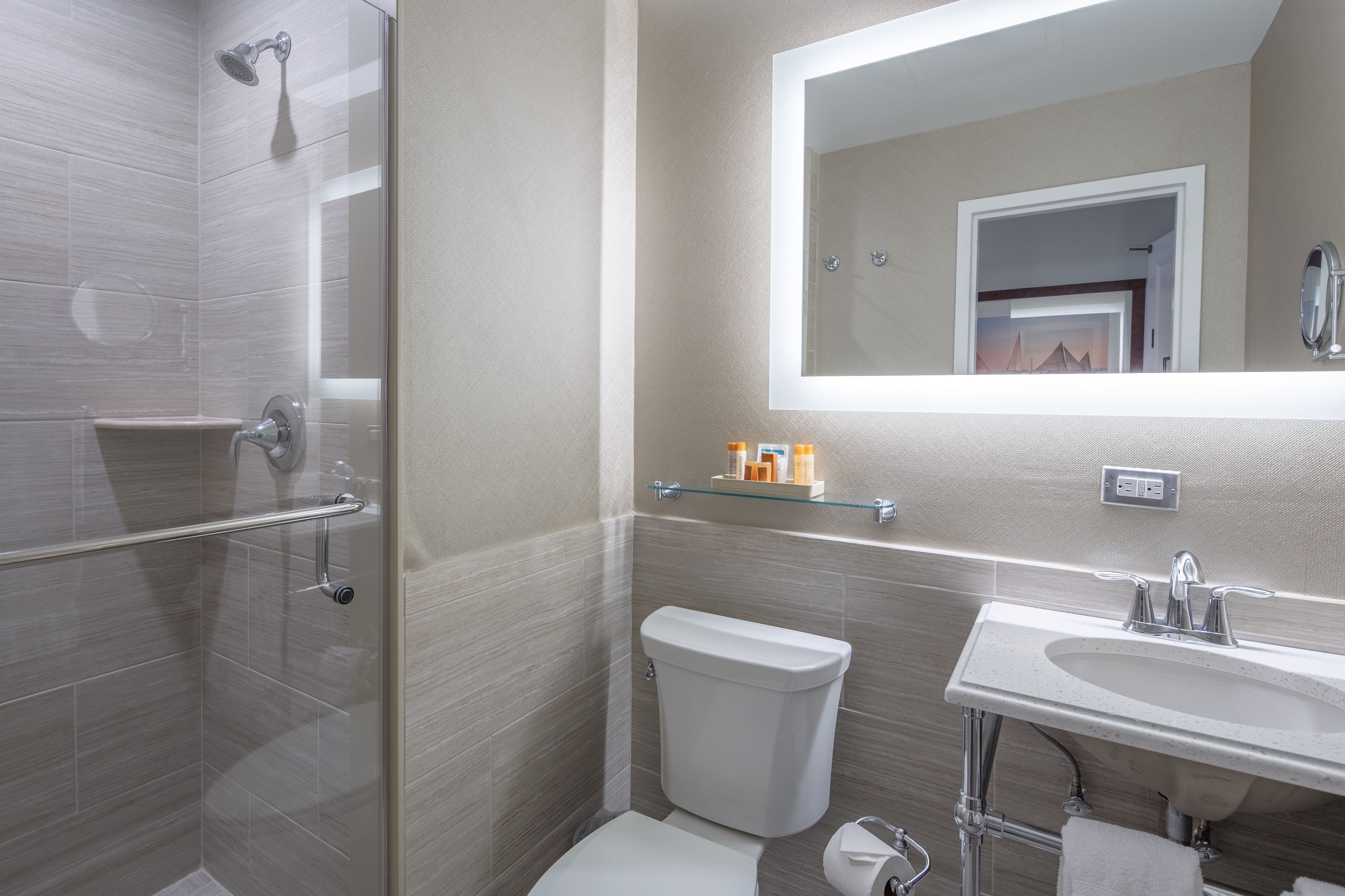 Un baño moderno con cabina de ducha de vidrio, inodoro, lavabo con espejo y un estante pequeño con artículos de aseo.