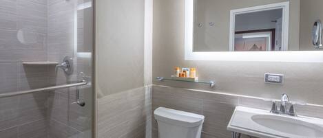 Un baño moderno con cabina de ducha de vidrio, inodoro, lavabo con espejo y un estante pequeño con artículos de aseo.
