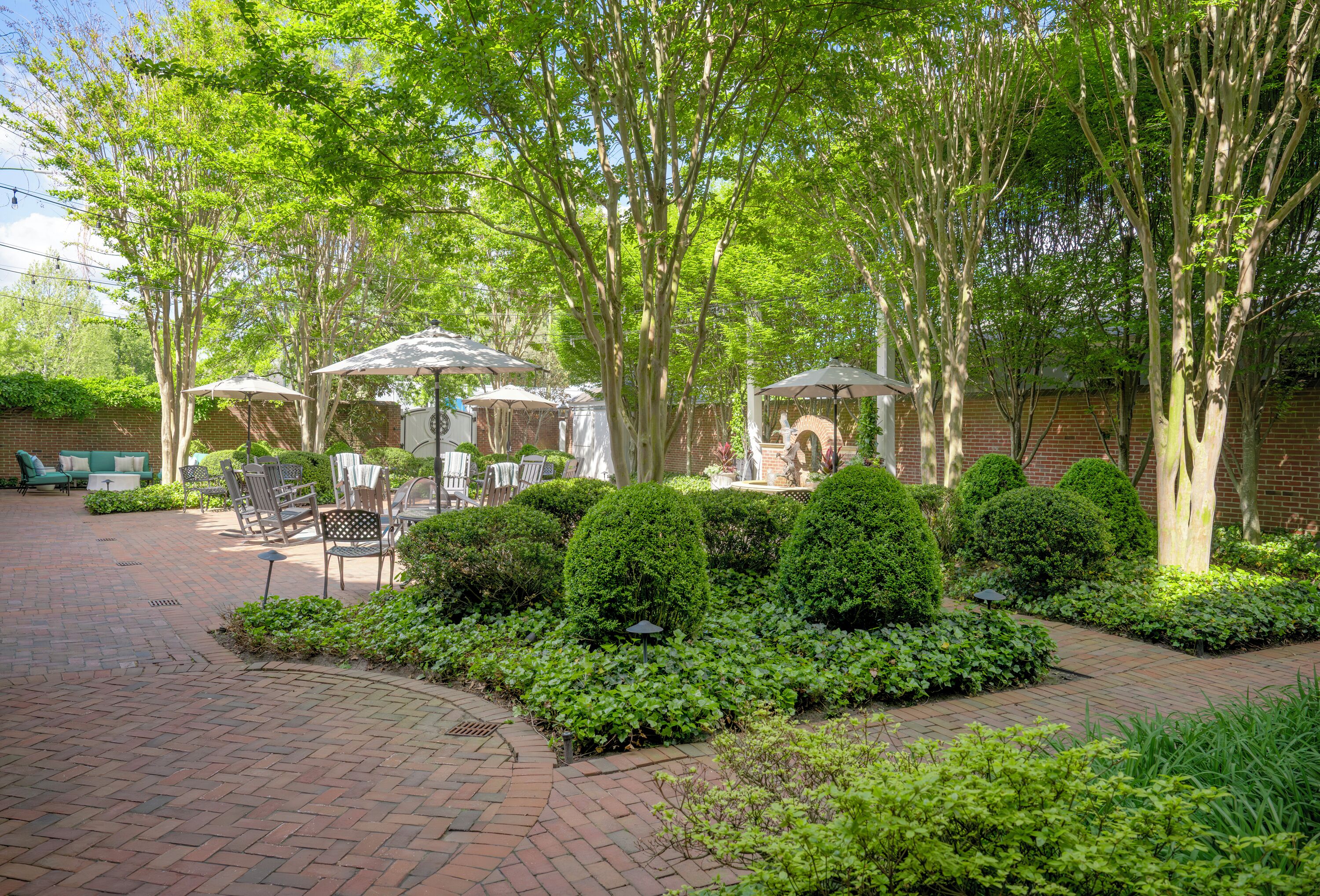 A courtyard with brick pathways, neatly trimmed bushes, and outdoor furniture under umbrellas.