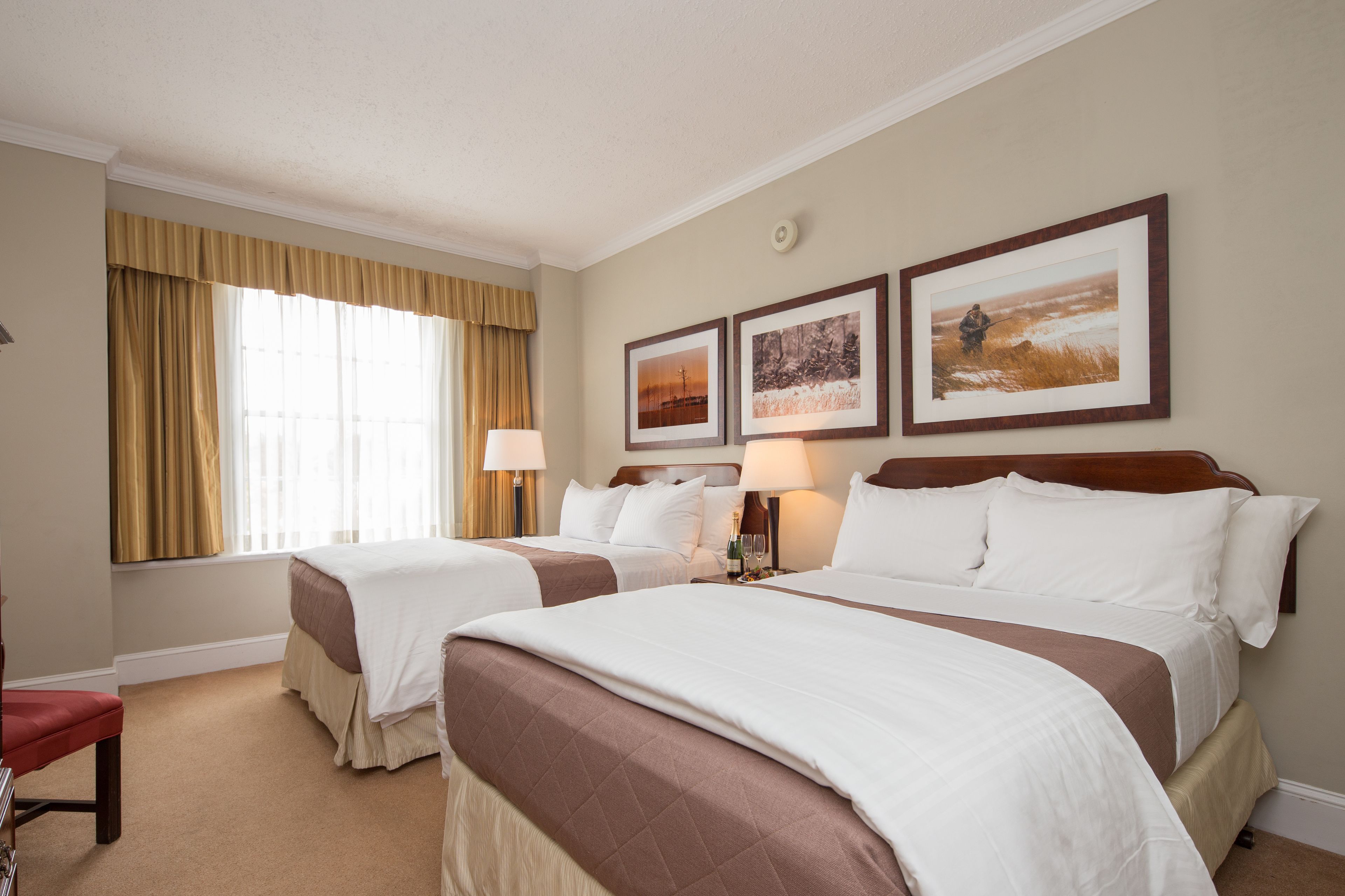 Una habitación de hotel con dos camas, un ventanal con cortinas y tres cuadros en la pared.
