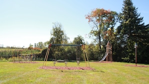 Une aire de jeux avec des balançoires, un toboggan et une structure d’escalade, située dans un espace verdoyant avec des arbres.