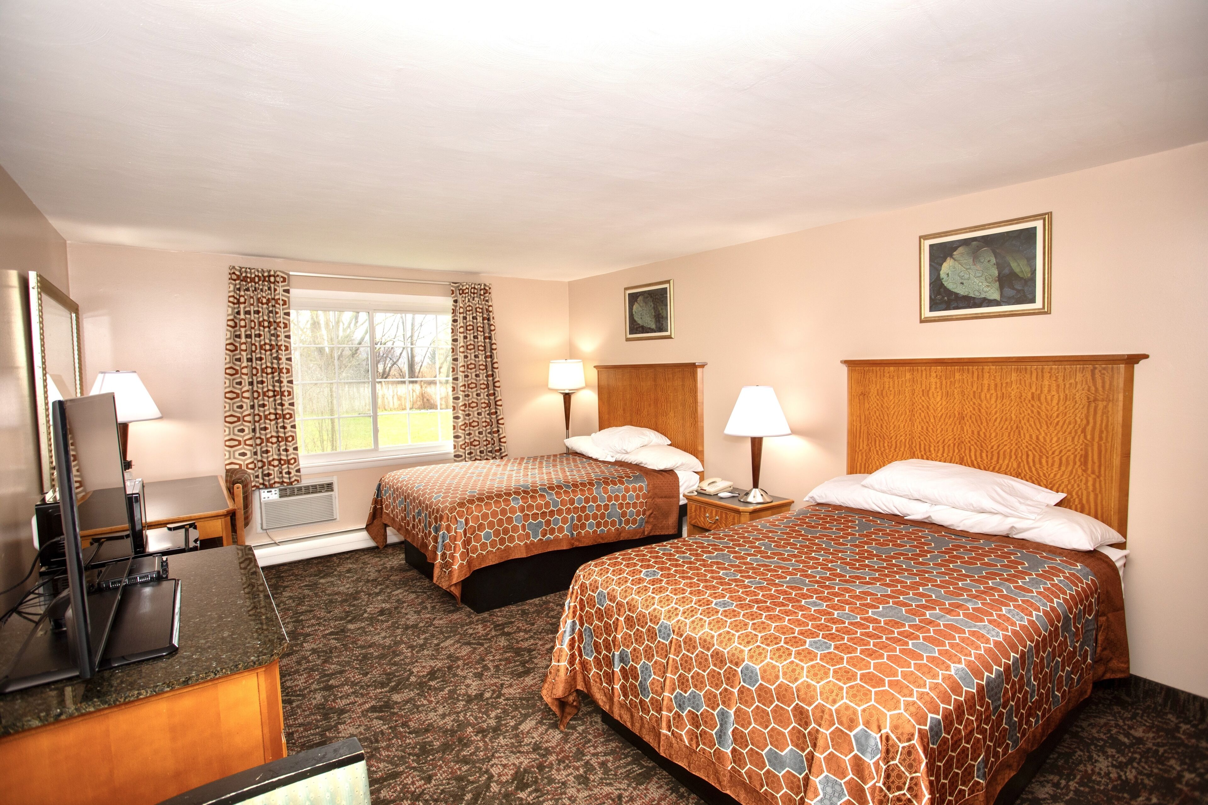 A hotel room with two beds, a desk with a computer, a window with patterned curtains, and a painting on the wall.
