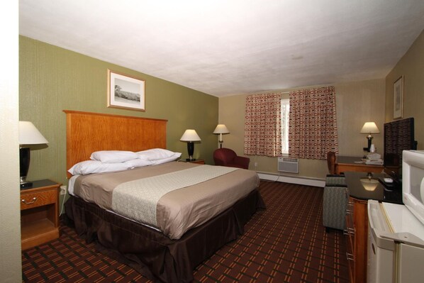 A hotel room with a bed, bedside lamps, a chair, a desk with a computer, a mirror, and a window with patterned curtains. - Budget Inn - Syracuse Airport (North Syracuse)