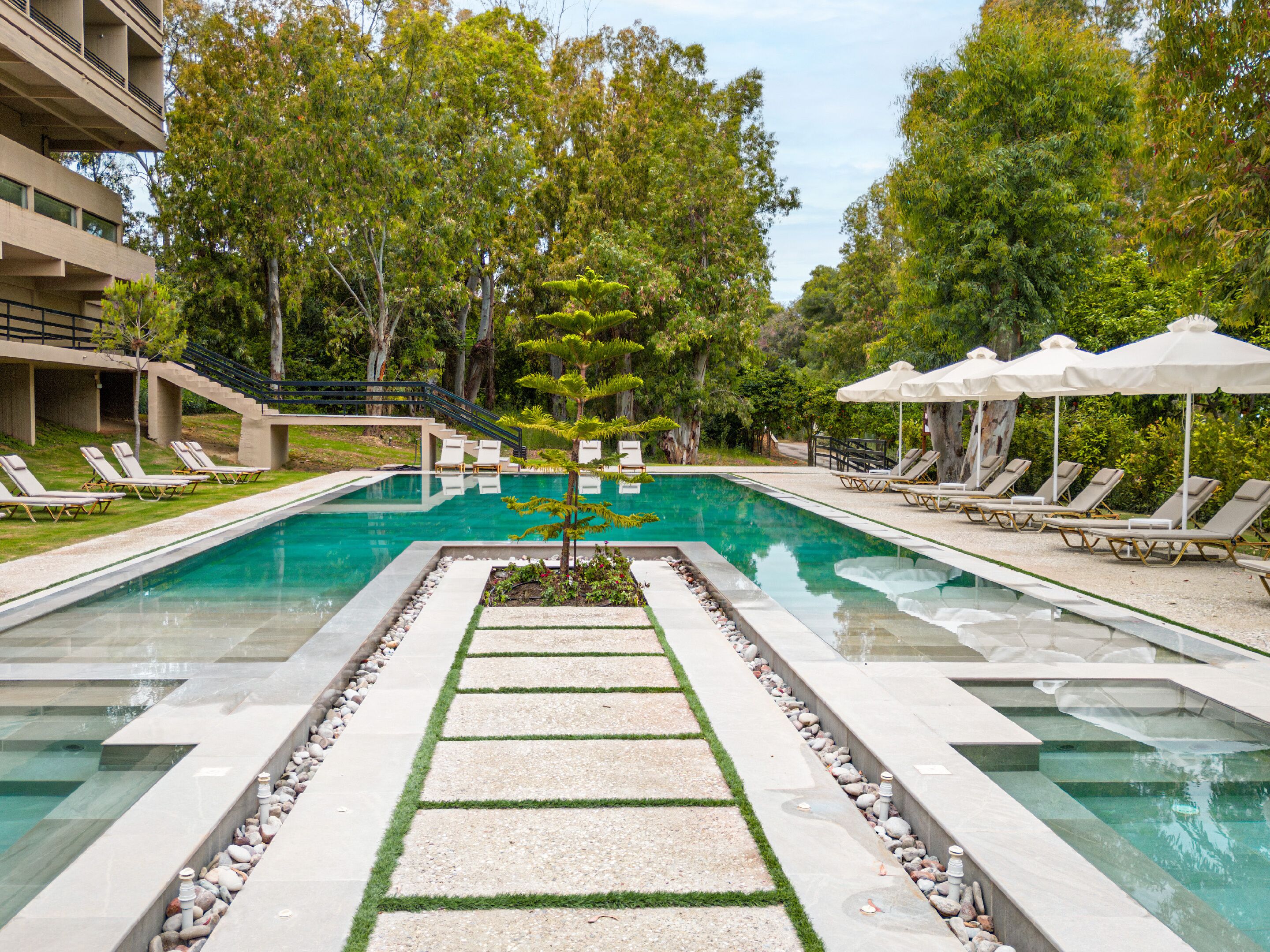 Une piscine entourée d’arbres et d’un bâtiment, avec des chaises longues et des parasols.