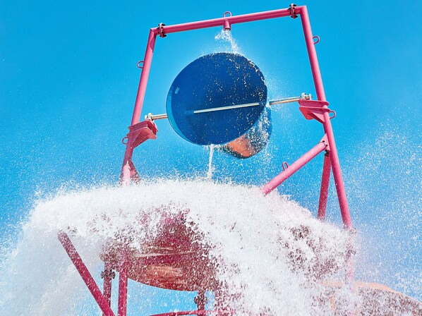 Una fuente de agua con cuenca circular y estructura de metal rojo, frente a un cielo azul despejado.