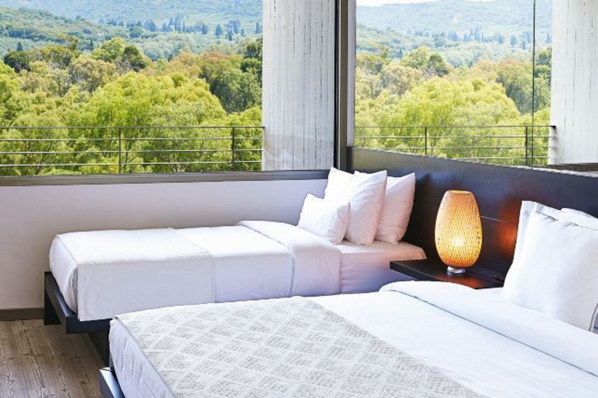 Une chambre d’hôtel avec deux lits, une table de chevet, une lampe et une vue sur un paysage verdoyant grâce à de grandes fenêtres.