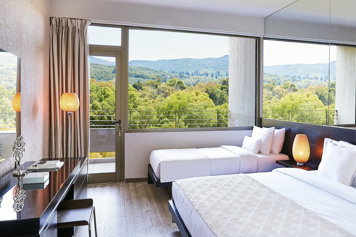 Habitación de hotel con balcón, dos camas, un escritorio y vistas a un paisaje boscoso.