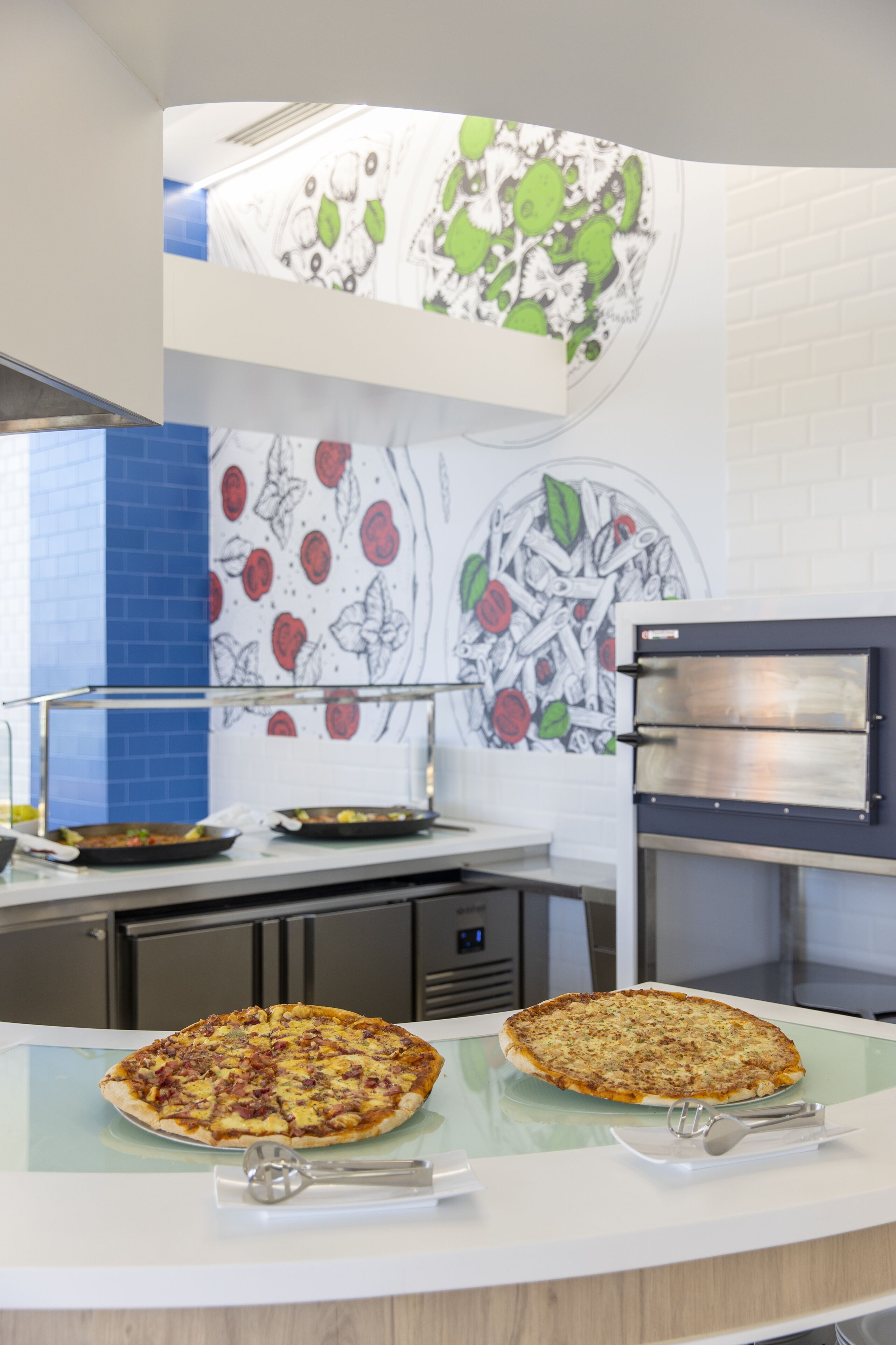 A kitchen with two pizzas on a glass counter, a pizza oven, and a wall with a fruit pattern.