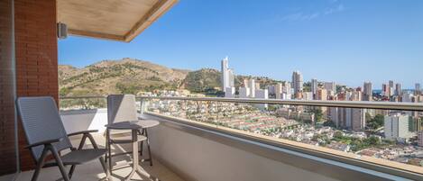Un balcón con dos sillas y una mesita pequeña, con vistas a un paisaje urbano que incluye edificios y colinas.