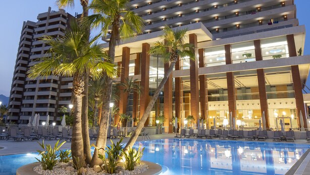 Un hôtel doté d’une piscine entourée de palmiers, d’une architecture moderne et offrant une vue sur un ciel dégagé.