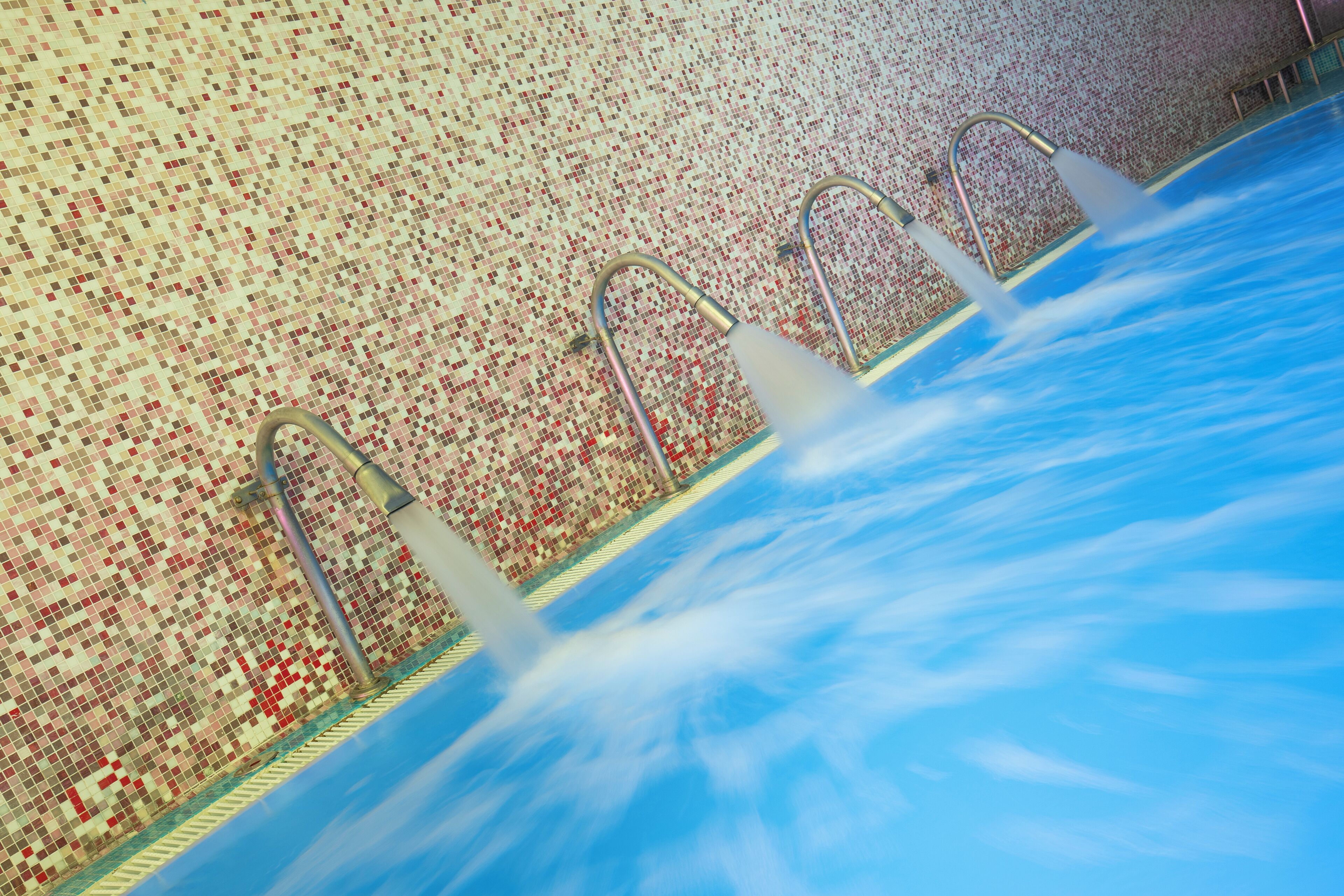 A swimming pool with three metal ladders and a mosaic tiled wall.