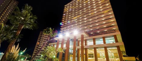 A modern hotel with a pool area, featuring a large pool, palm trees, and a well-lit facade.