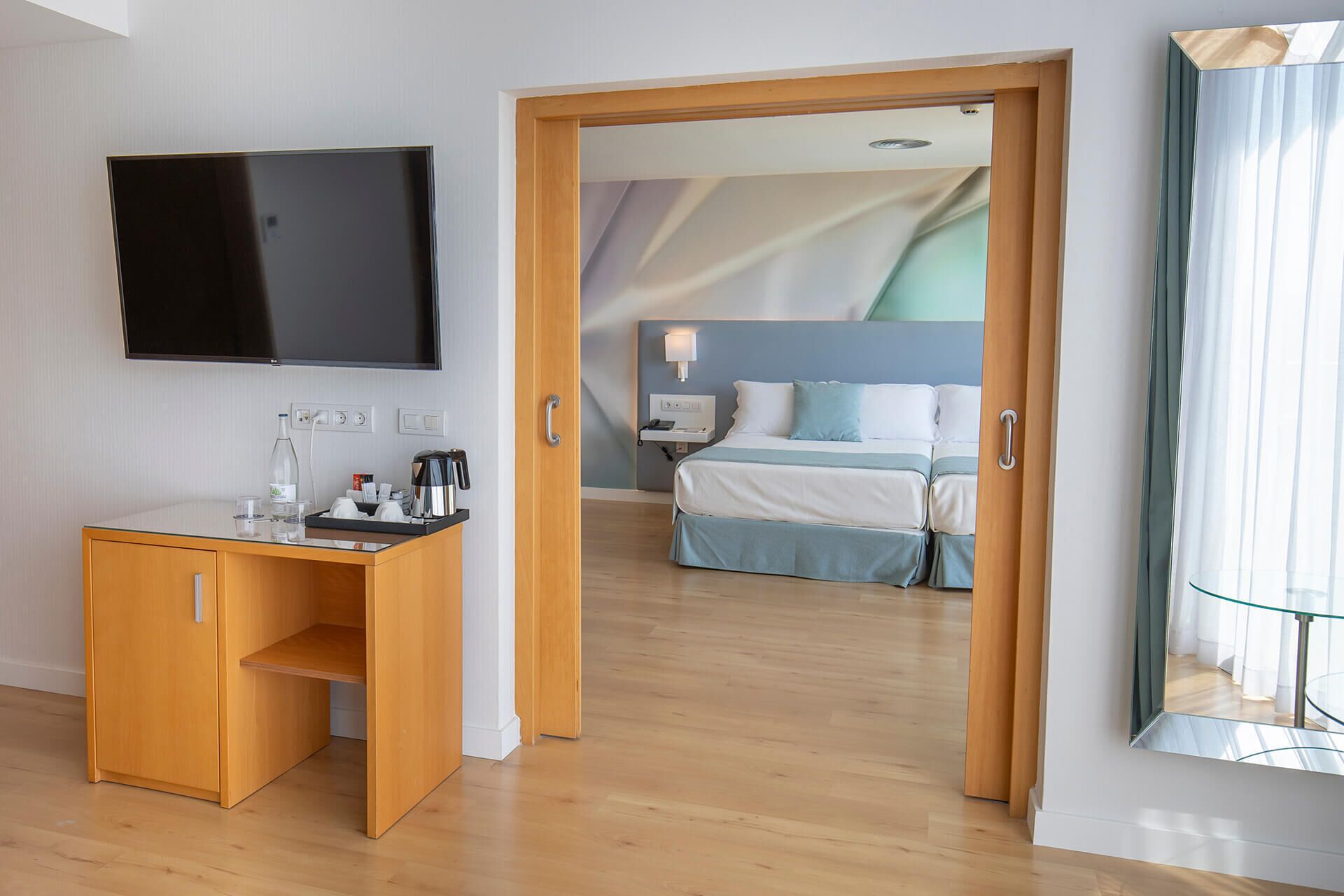 A hotel room with a wooden desk, a flat-screen TV, a bed with a blue headboard, and a glass partition.