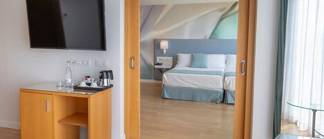 A hotel room with a wooden desk, a flat-screen TV, a bed with a blue headboard, and a glass partition.