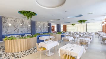 Un restaurante moderno con mesas y sillas blancas, piso estampado y plantas verdes en un macetero de madera.
