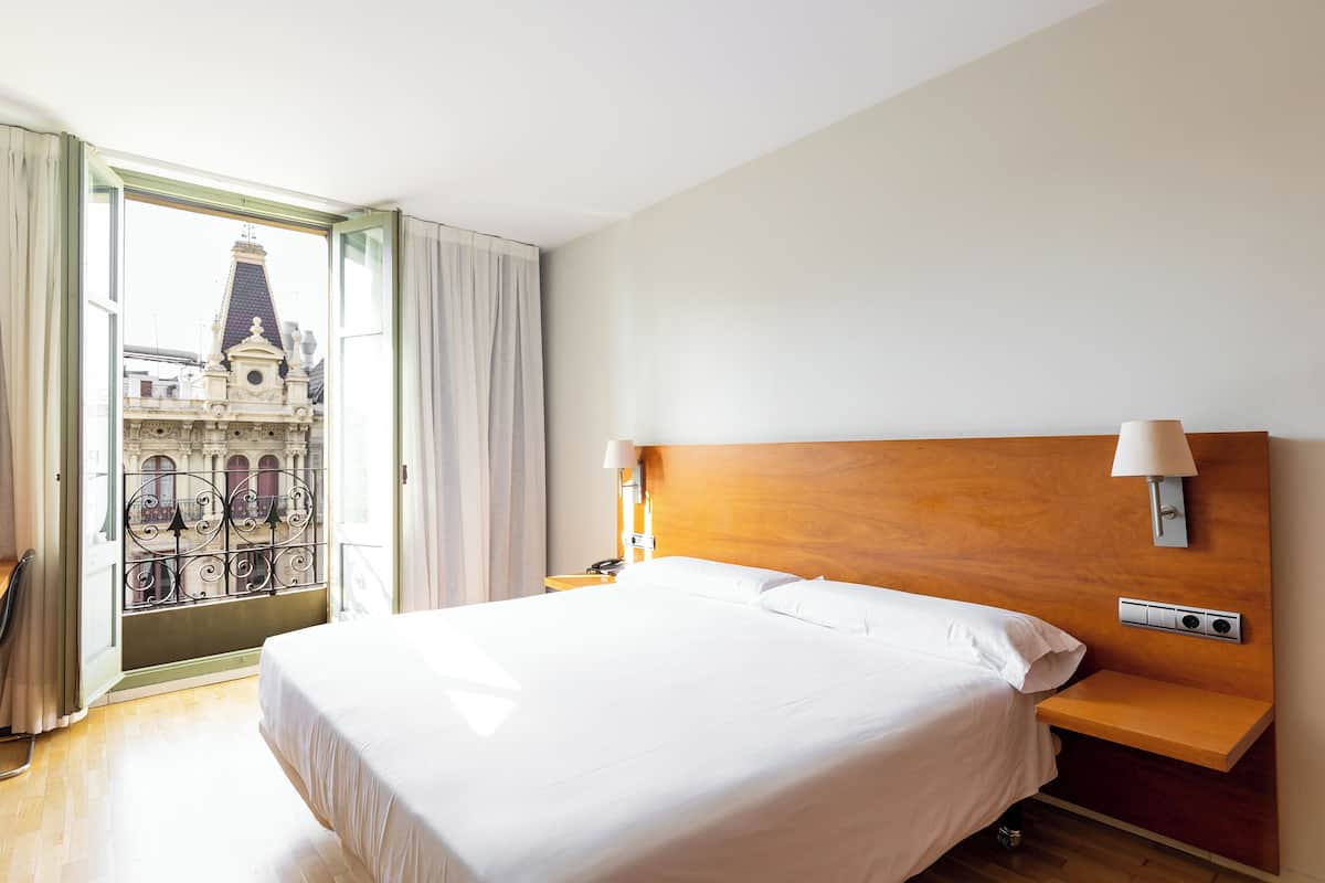 Habitación de hotel con una cama grande, cabecera de madera, lámparas de noche y vista a un edificio a través de una ventana abierta.