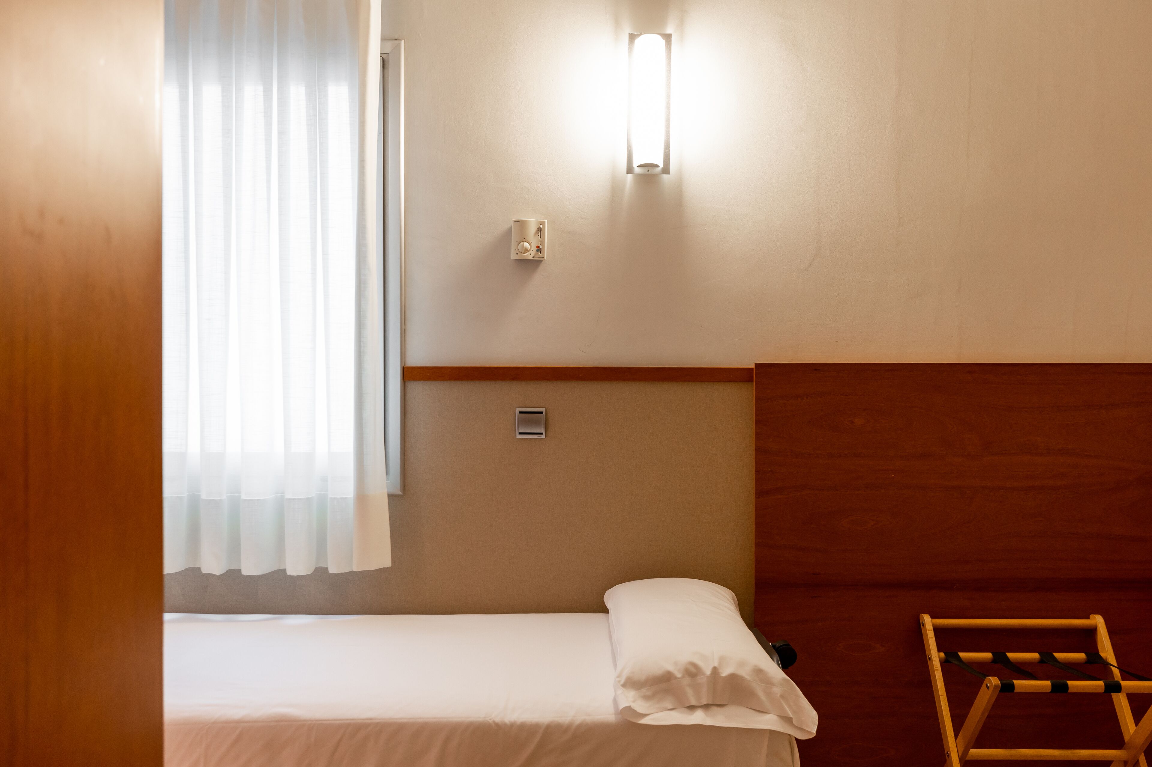Un llit individual amb capçalera de fusta, una taula de nit, una làmpada penjada a la paret i una finestra amb cortines translúcides.