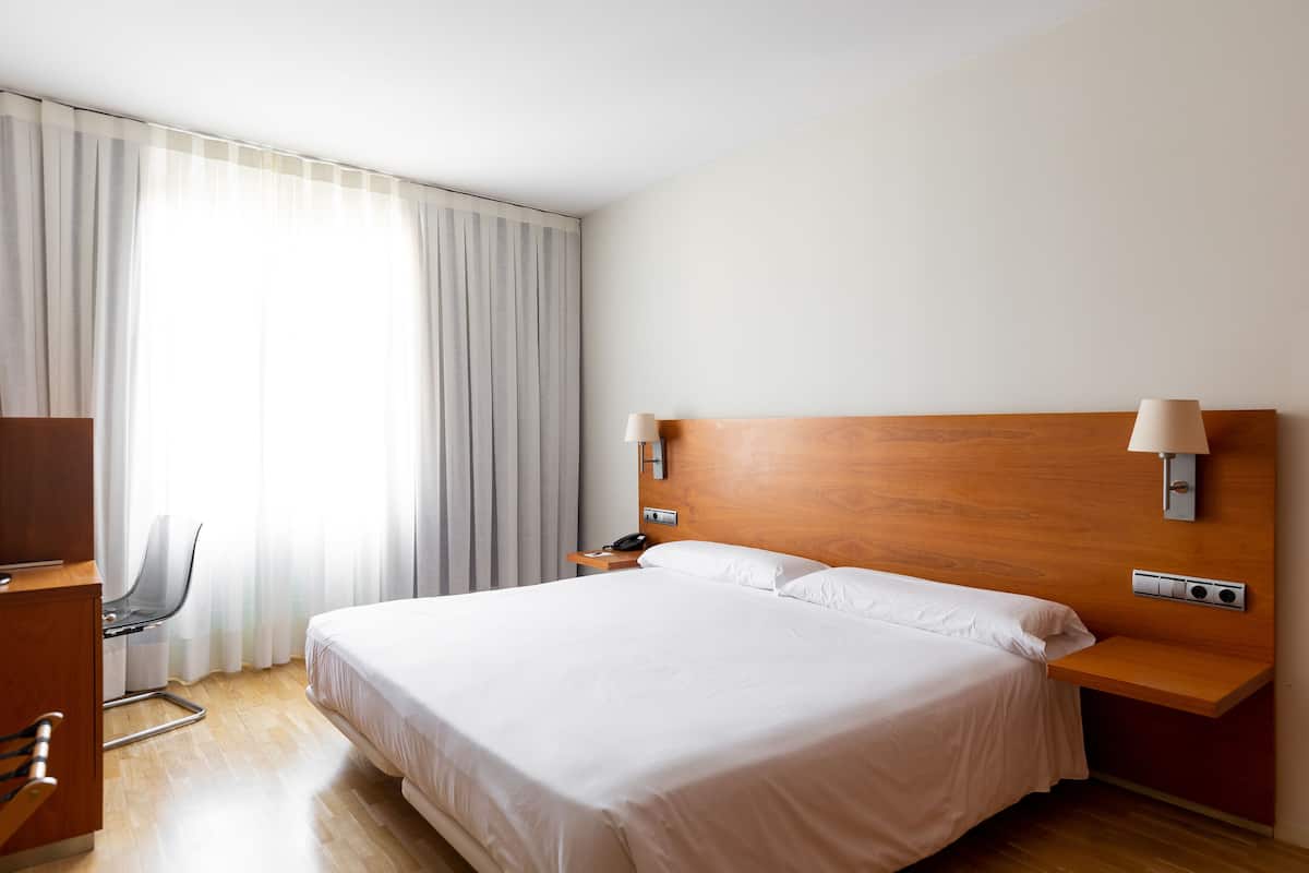 Una habitación de hotel con una cama grande, un escritorio con silla, una mesita de noche y una lámpara de pared.