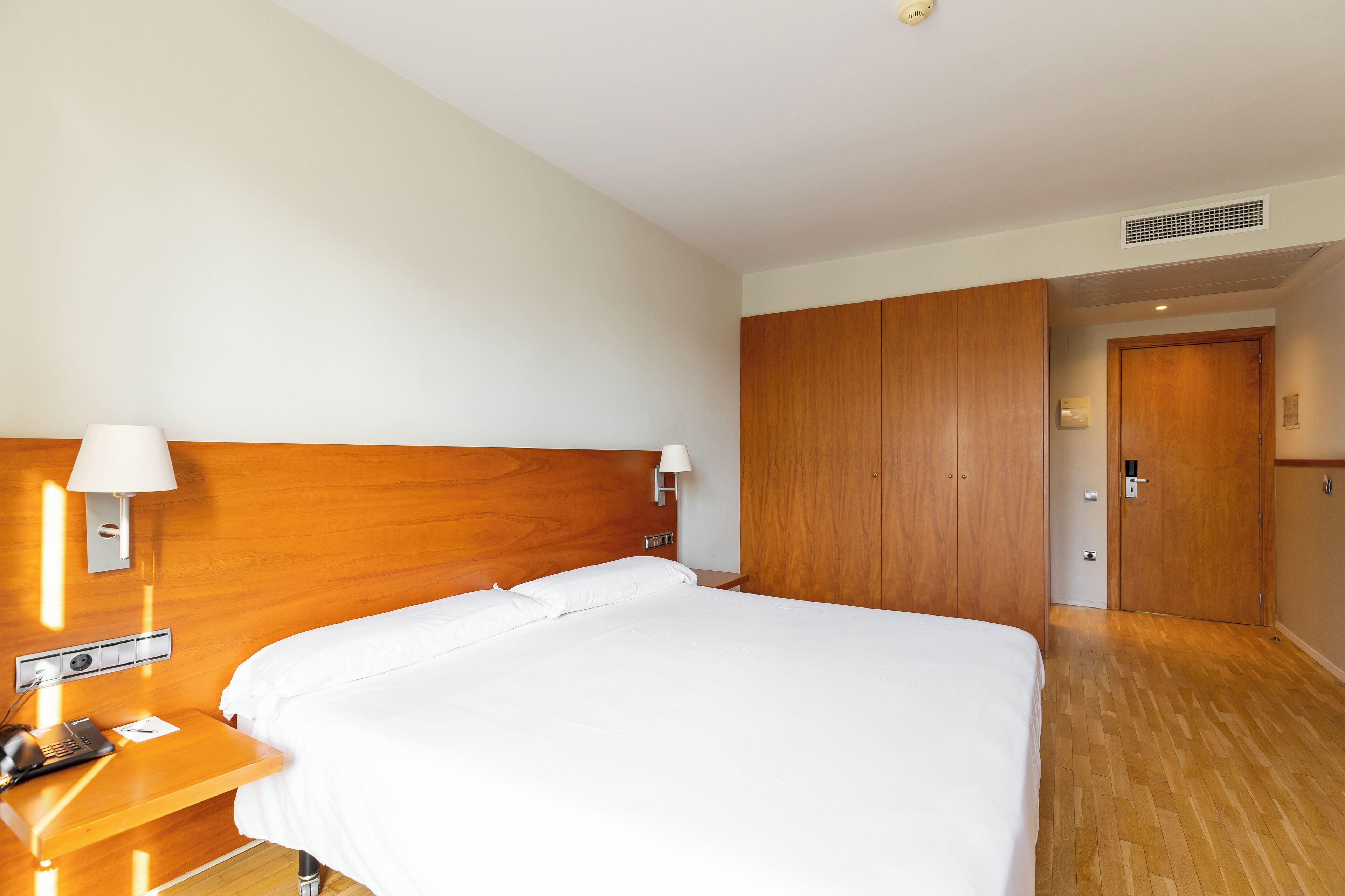 Habitación de hotel con cabecera de madera, una cama individual, una mesita de noche con lámpara y una puerta que da a otra habitación.