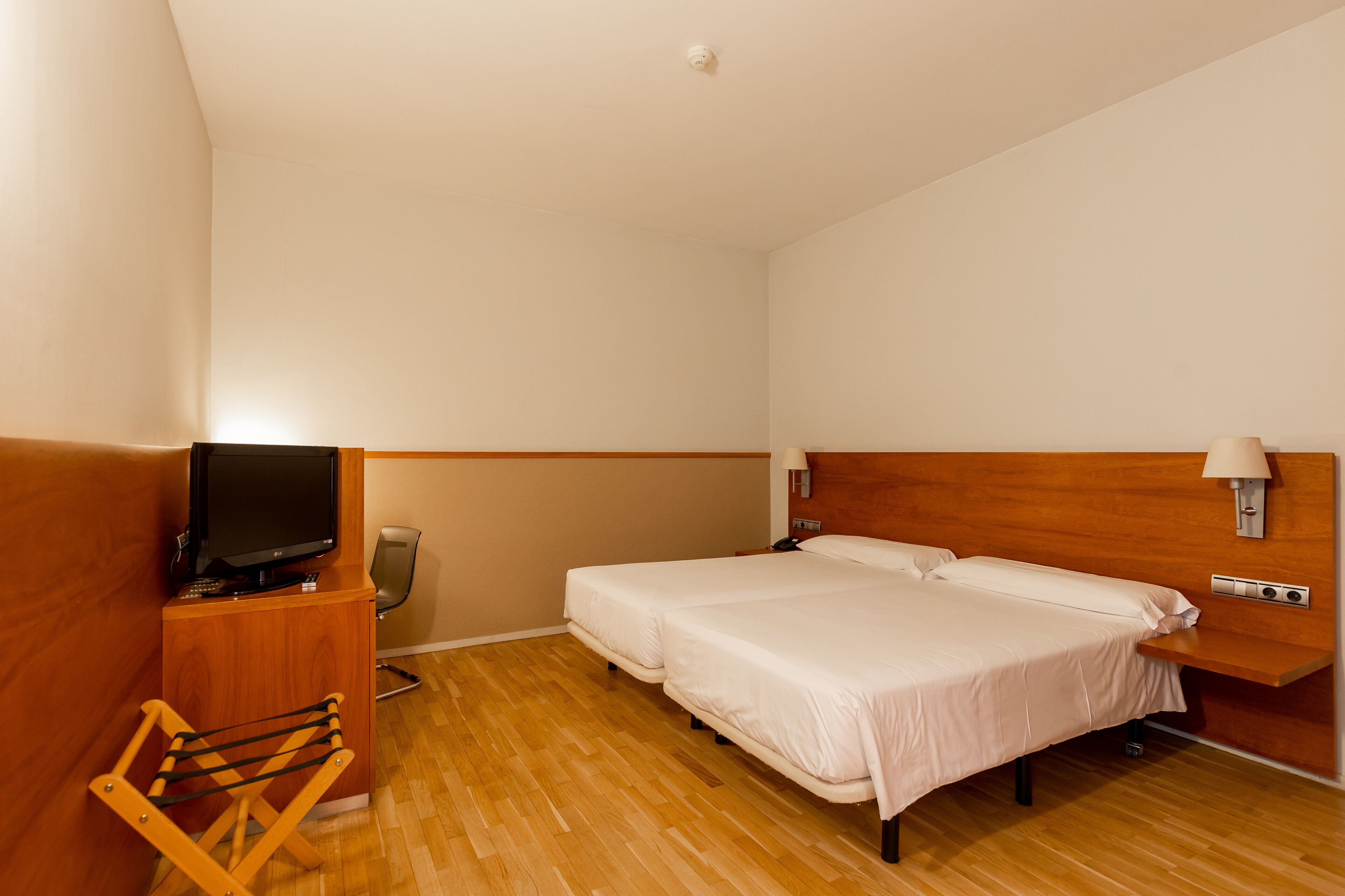 A hotel room with a wooden floor, two single beds, a TV on a wooden stand, a chair, a desk with a lamp, and a wall-mounted light fixture.