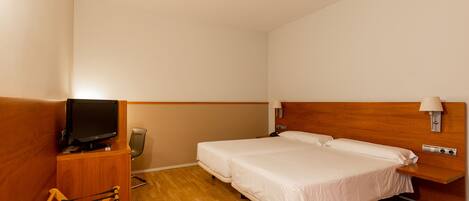 A hotel room with a wooden floor, two single beds, a TV on a wooden stand, a chair, a desk with a lamp, and a wall-mounted light fixture.