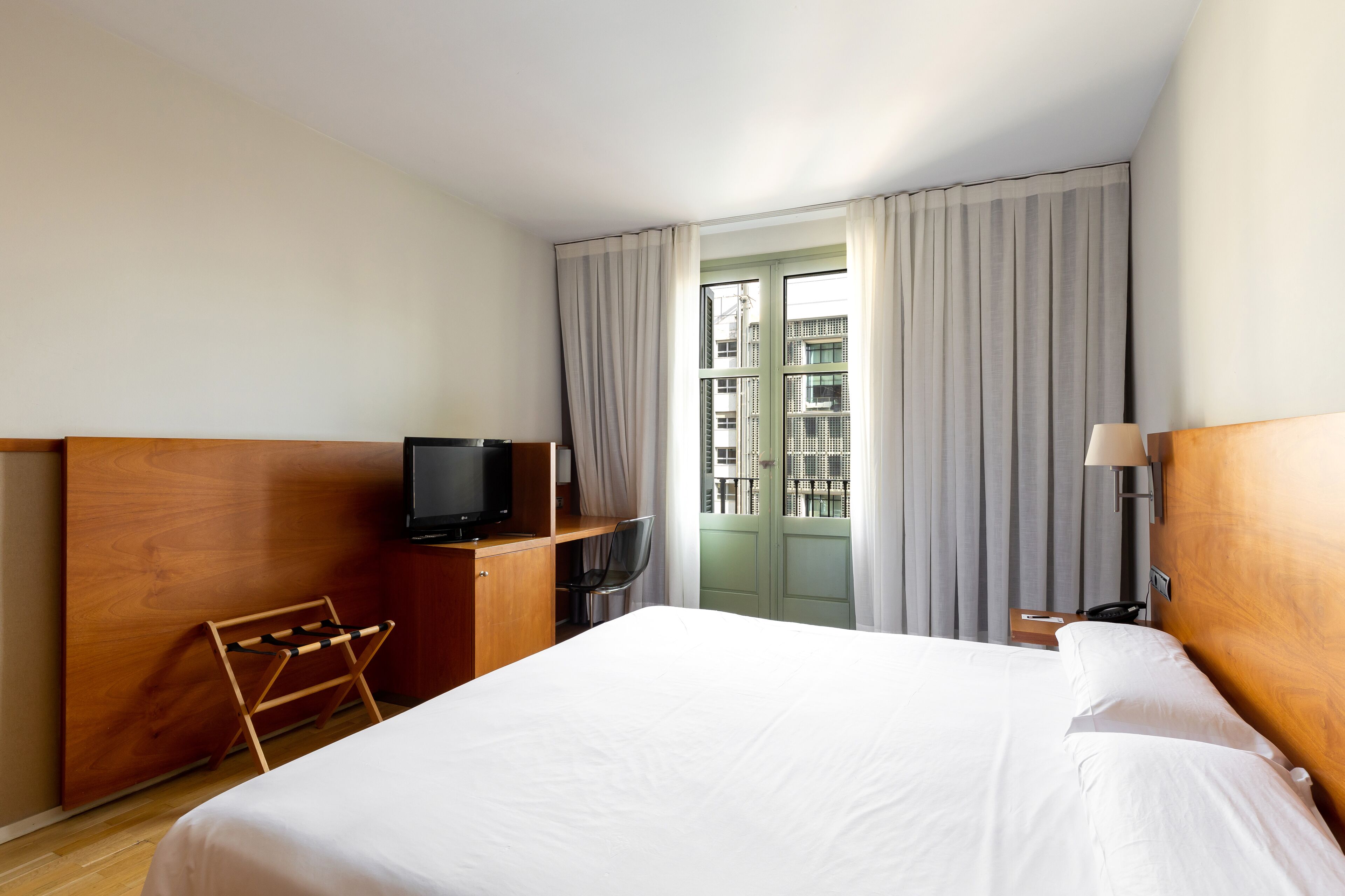 Habitación de hotel con una cama grande, cabecera de madera, un televisor sobre una estantería de madera, un escritorio con una silla y vistas a edificios a través de una ventana con cortinas.