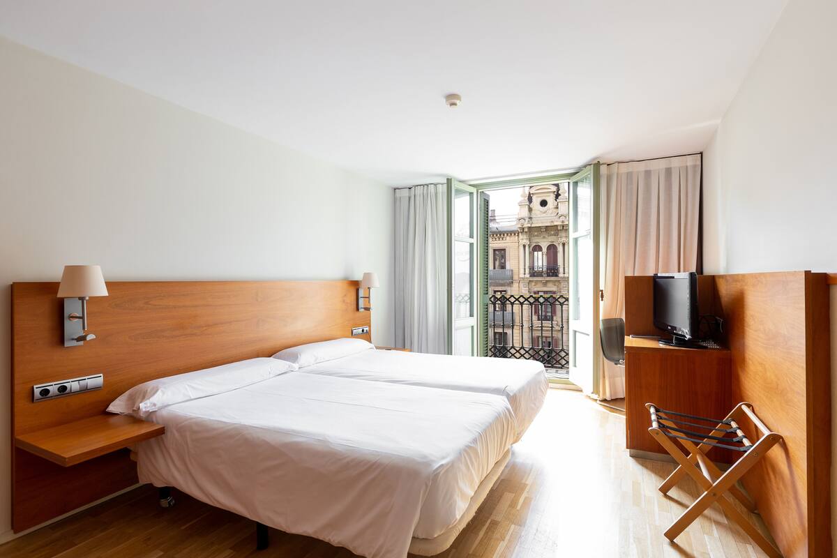 Habitación de hotel con cama doble, televisión, mesita de noche de madera y balcón con vista a un edificio.