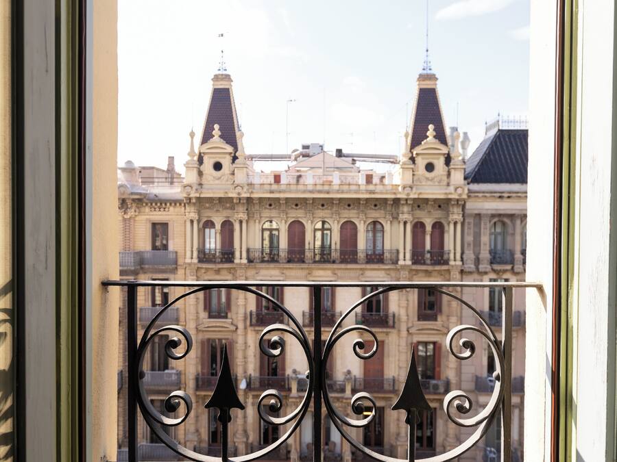 Vista a través de una ventana con barandillas de hierro ornamentado, mostrando un edificio histórico con dos torres destacadas y balcones.