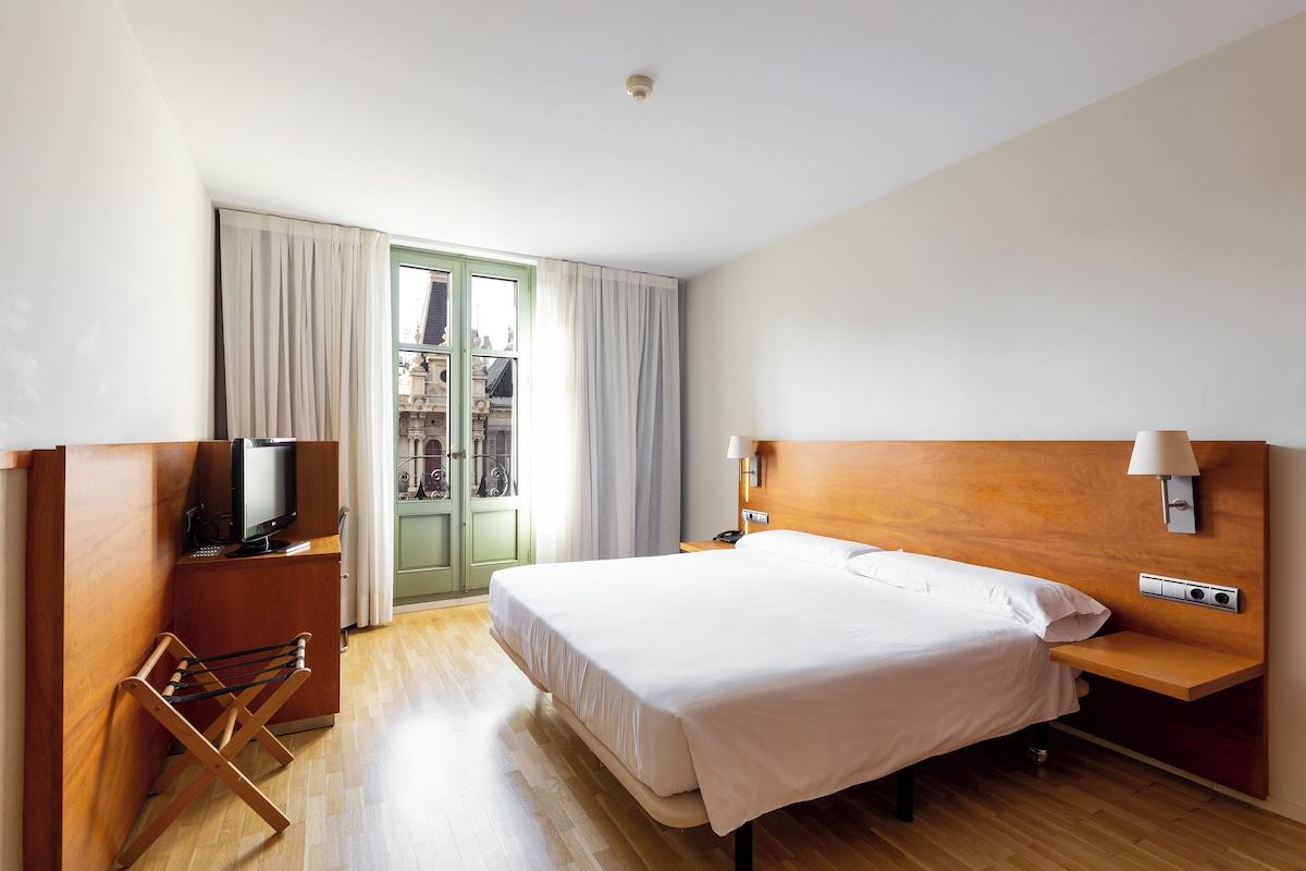 A hotel room with a bed, a TV, a wooden nightstand, a lamp, and a view of the outside.
