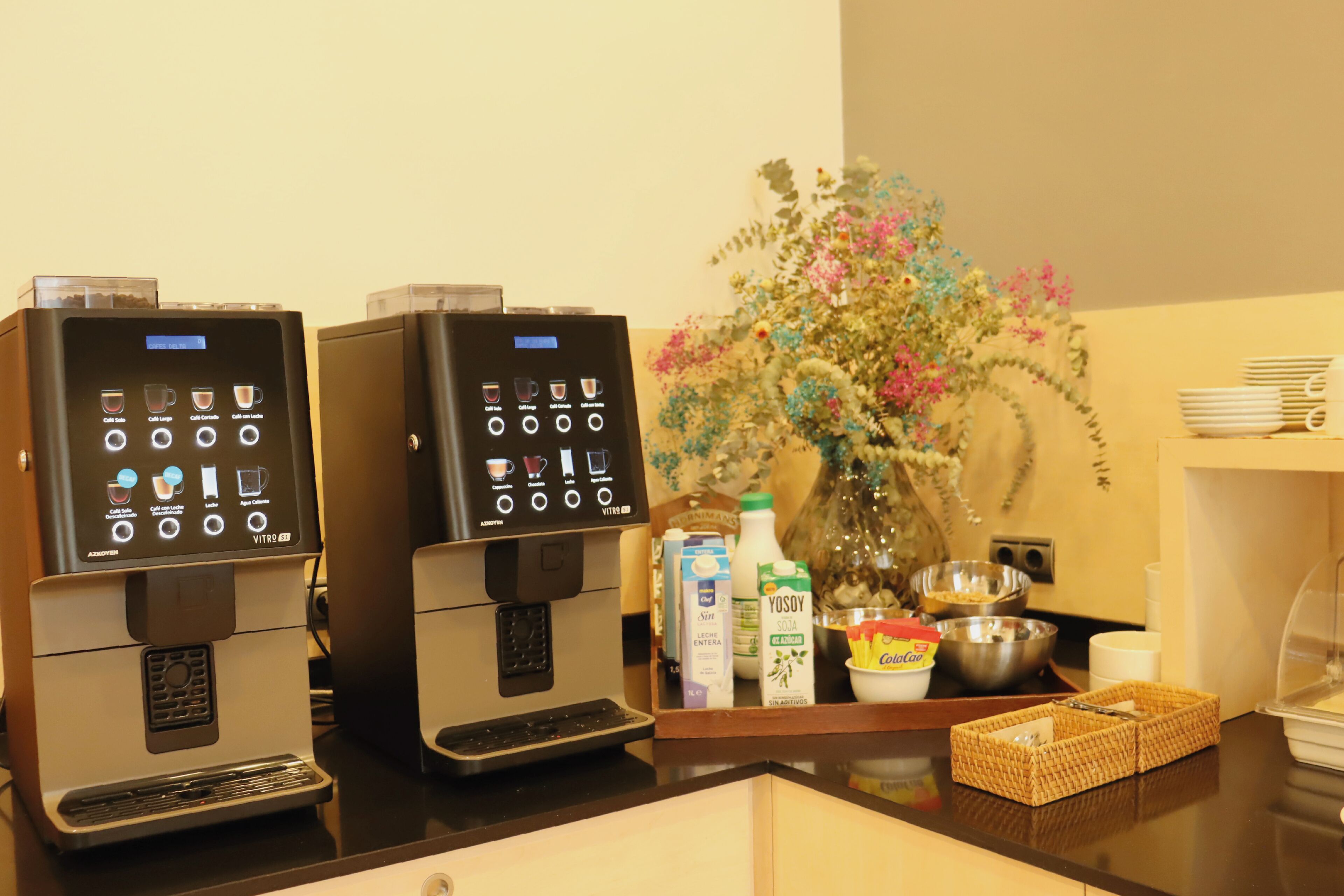 A coffee station with two espresso machines, a variety of coffee pods, milk and soy milk cartons, a bowl of cereal, and a basket of utensils.