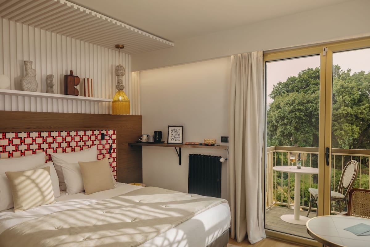A bedroom with a bed, a bedside table, a radiator, a window with curtains, and a balcony with a table and chairs.