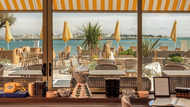 Een terras aan zee met rieten meubilair, gestreepte parasols en uitzicht op zee en de stad.