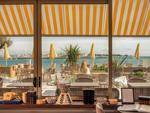 A seaside terrace with wicker furniture, striped umbrellas, and a view of the sea and cityscape. - Hôtel Les Brises (La Rochelle)