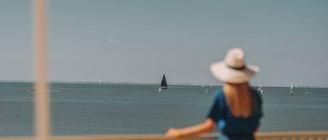 Une personne vêtue d’une robe bleue et d’un chapeau blanc, debout et contemplant une mer calme où voguent des voiliers, à l’ombre d’un parasol rayé.