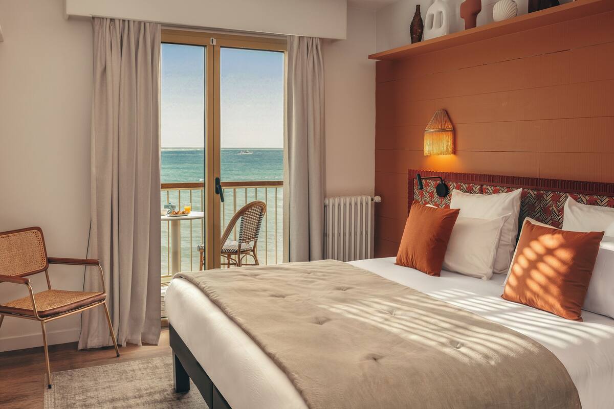 A hotel room with a bed, a chair, a view of the sea, and a shelf with decorative items.