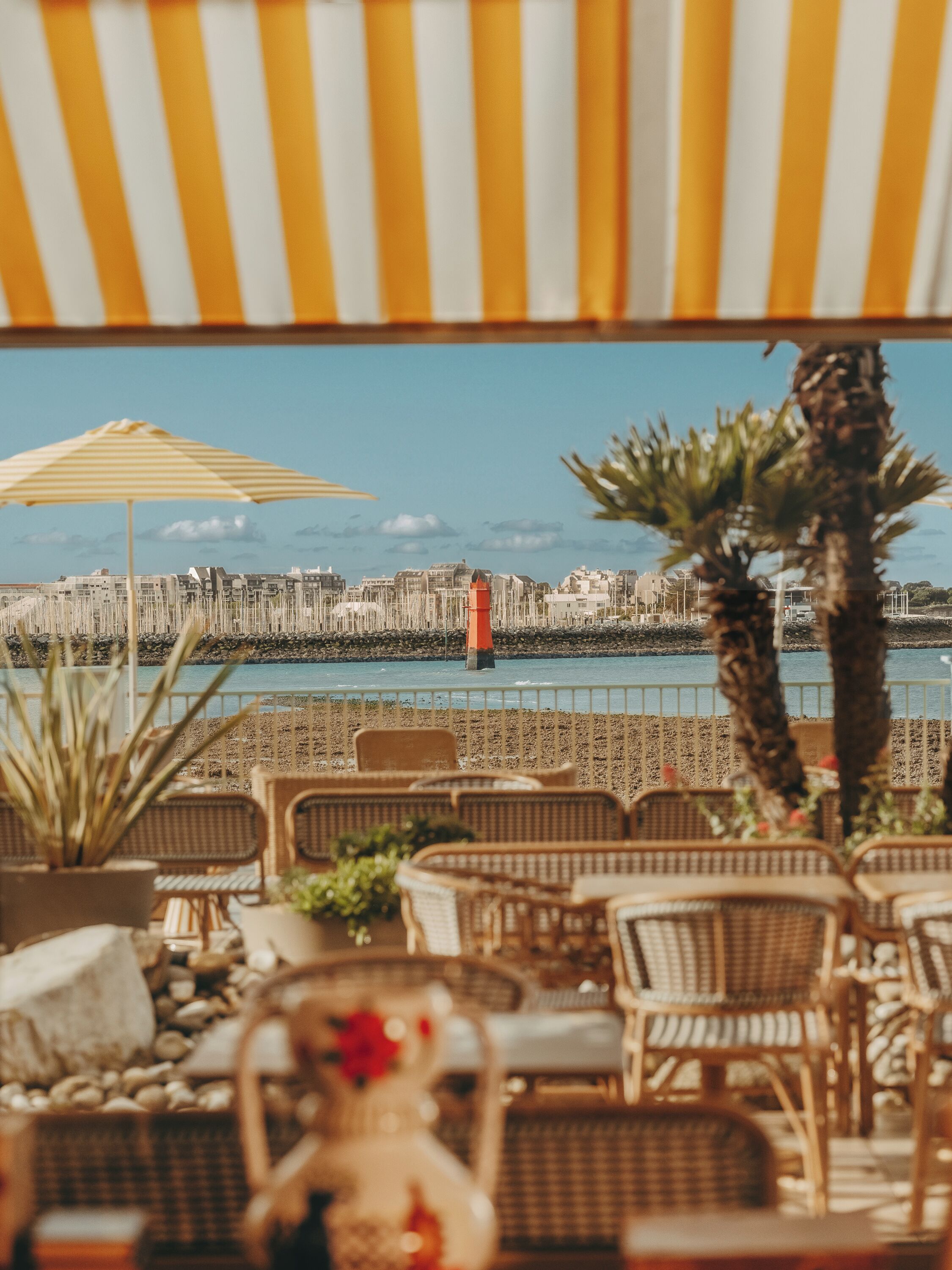 A seaside terrace with wicker furniture, a striped umbrella, and a view of a cityscape across the water.