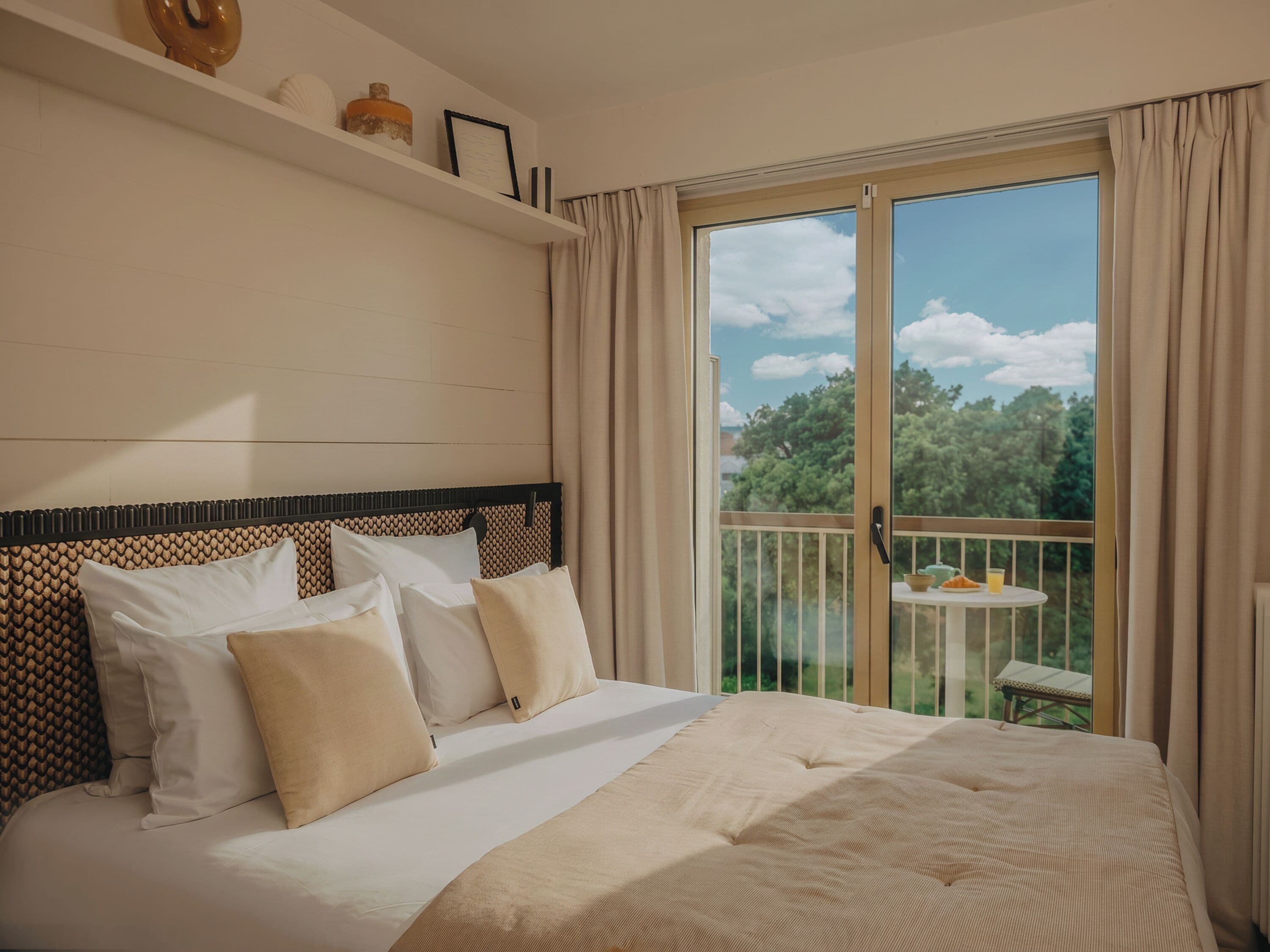 A hotel room with a large bed, a balcony with a table and chairs, and a view of trees.