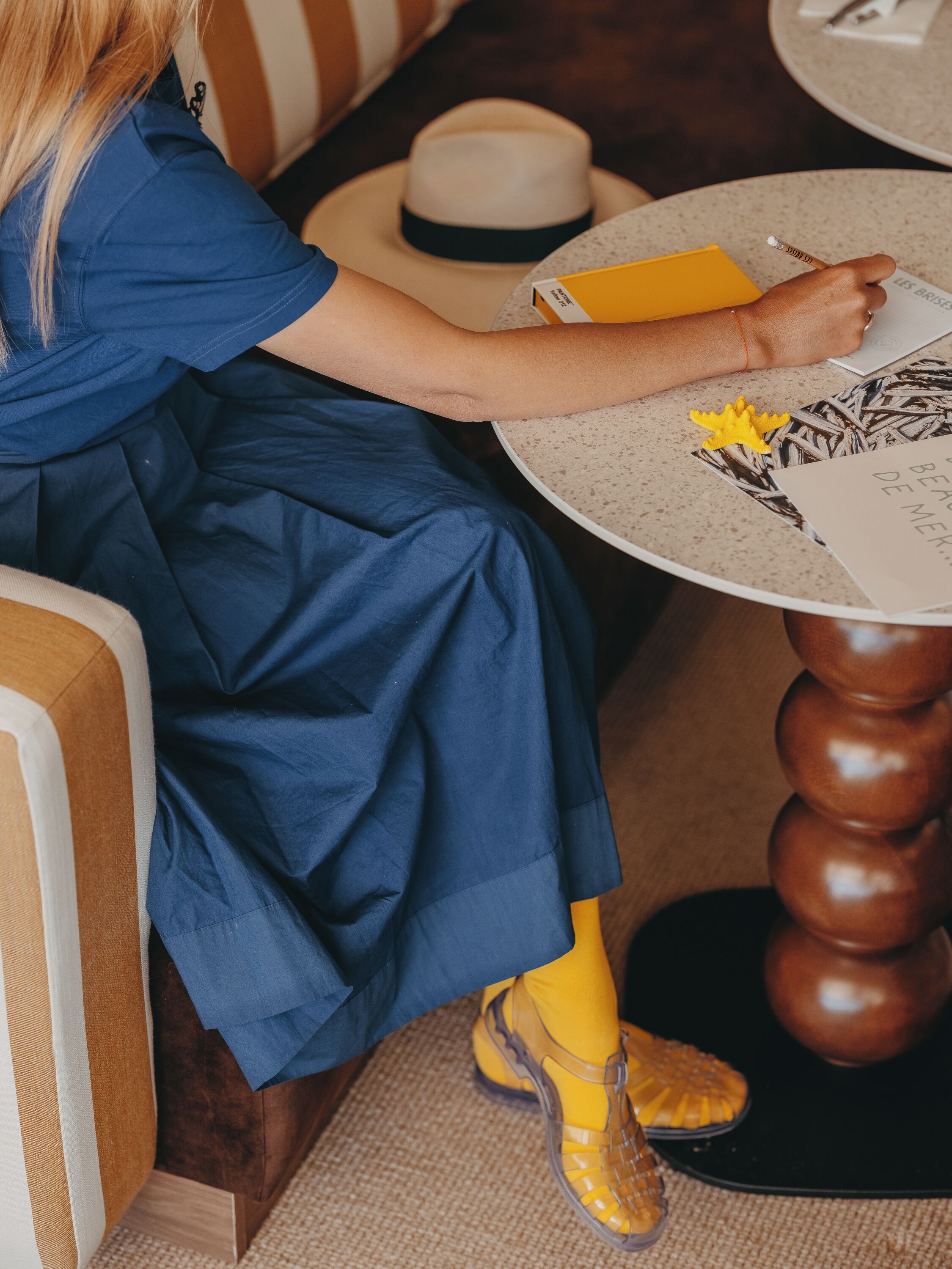 Una persona seduta a un tavolo rotondo con un cappello, un quaderno giallo e un libro.