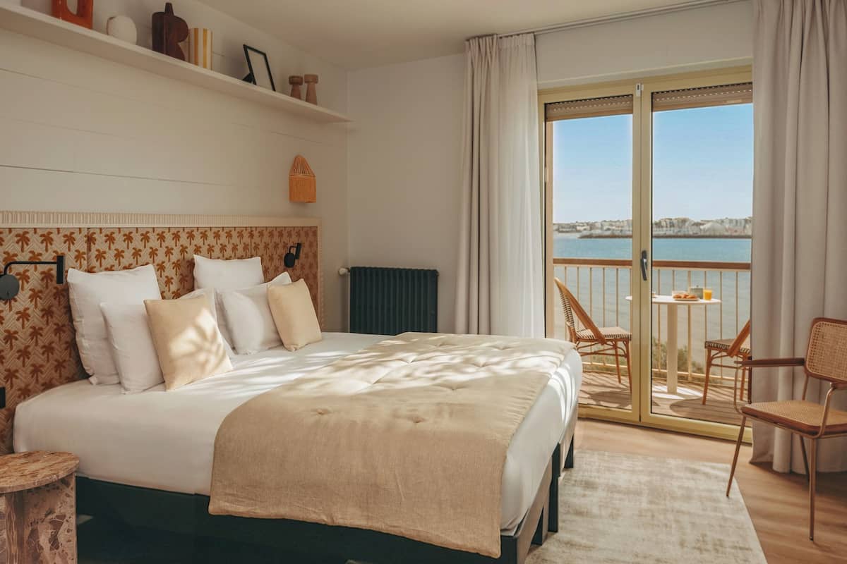 A hotel room with a bed, a chair, a table with a drink, and a view of the sea.