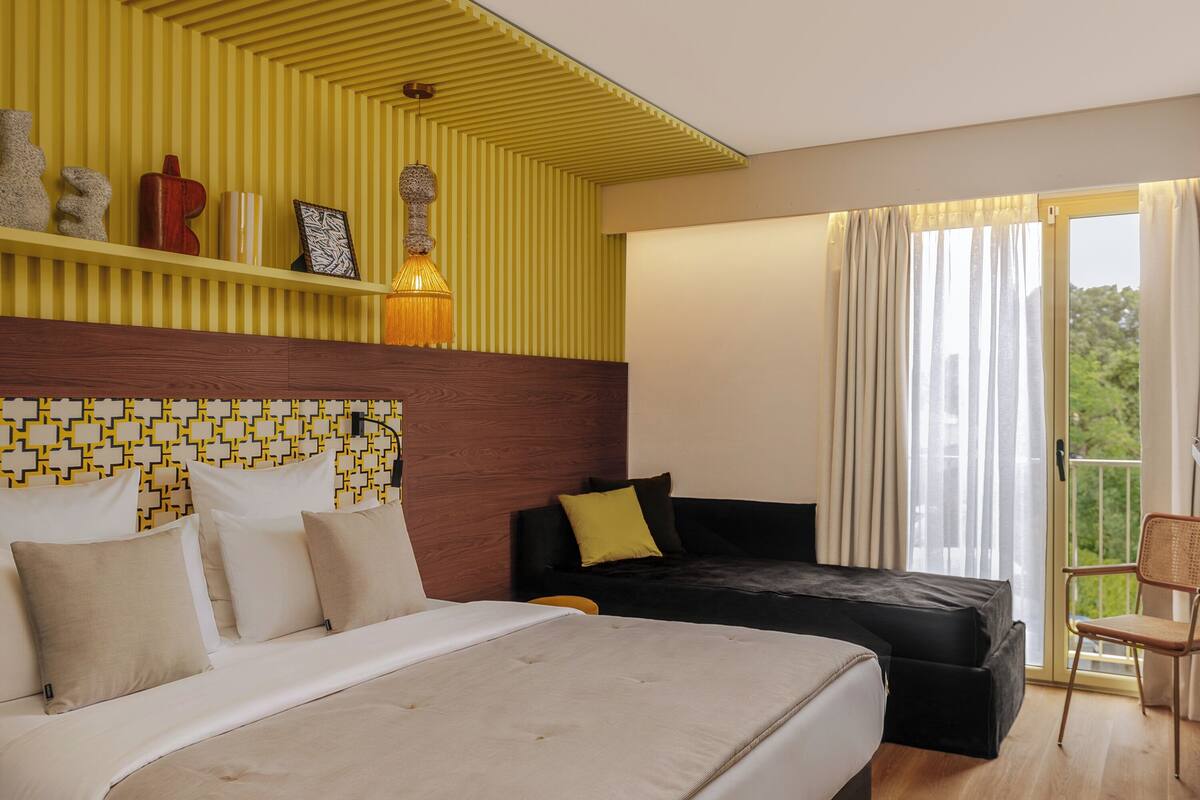 A hotel room with a large bed, a smaller sofa, a wooden headboard, a yellow wall with a geometric pattern, a window with curtains, and a balcony with a chair.