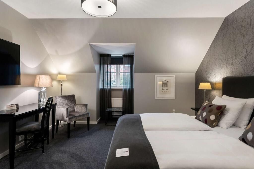 A hotel room with a bed, a desk, a chair, a lamp, and a window.