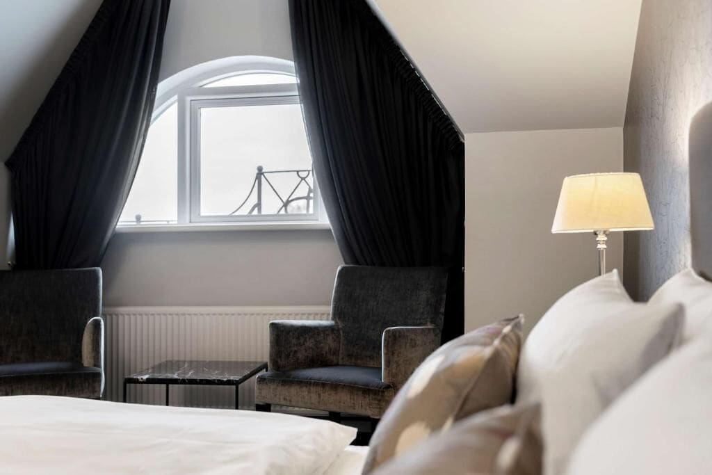 Une chambre d’hôtel avec un lit, deux fauteuils, une petite table, une fenêtre avec des rideaux et une lampe.